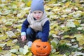 Happy little boy in cap and vest is sitting with halloween pumpkin surrounded by fallen leaves.