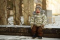 Portrait of happy little boy with red cheeks, in bright clothes and mittens sitting on bench in snowy winter park Royalty Free Stock Photo