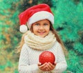 Portrait happy little girl child in santa red hat holding ball toy outdoors over a christmas tree Royalty Free Stock Photo