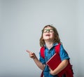 Portrait of happy little girl in glasses hugging a book. Child with backpack laughing and pointing with finger up Royalty Free Stock Photo