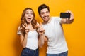Portrait of happy man and woman taking selfie photo on smartphone while showing peace sign, isolated over yellow background Royalty Free Stock Photo