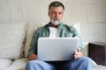 Portrait of happy mature man in casual clothes using laptop lying on sofa in house. Royalty Free Stock Photo