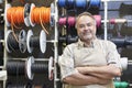 Portrait of a happy mature salesperson standing in front of electrical wire spool with arms crossed in hardware store