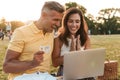 Portrait of happy middle-aged couple holding credit card while using laptop computer during picnic in summer park Royalty Free Stock Photo