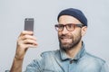 Portrait of happy middle aged smiling male wears denim jacket, hat and glasses, holds smart phone for making selfie, isolated over Royalty Free Stock Photo