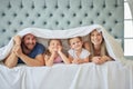 Portrait of happy parents with little children lying on bed at home under blanket. Caucasian girls bonding with their Royalty Free Stock Photo