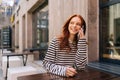 Portrait of happy red-haired young woman talking on mobile phone sitting at table in outdoor cafe terrace in summer day. Royalty Free Stock Photo