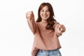 Portrait of happy redhead girl choosing you, pointing fingers at camera and smiling, inviting to event, beckon to join