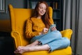 Portrait of happy redhead young woman using smartphone sitting in yellow armchair, smiling looking to device screen. Royalty Free Stock Photo