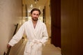 Portrait of a happy relaxed handsome middle aged European man dressed in white terry bathrobe standing on the hallway of a luxury