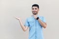 Portrait of happy satisfied handsome young bearded man in blue shirt standing, holding, pointing something and looking at camera