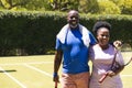 Portrait of happy senior african american couple with tennis rackets embracing on sunny grass court Royalty Free Stock Photo