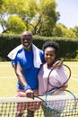 Portrait of happy senior african american couple with tennis rackets embracing on sunny grass court Royalty Free Stock Photo