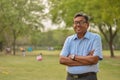 Portrait of a happy senior Indian man wearing a shirt and hands crossed in the outside setting standing and thinking in a park in Royalty Free Stock Photo