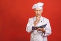 Portrait of a happy senior male chef cook holding recipe book and preparing food isolated on a red background Royalty Free Stock Photo