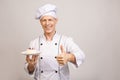 Portrait of a happy senior male chef dressed in uniform holding plate with piece of cake and looking at camera isolated over gray Royalty Free Stock Photo