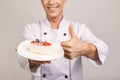 Portrait of a happy senior male chef dressed in uniform holding plate with piece of cake and looking at camera  over gray Royalty Free Stock Photo