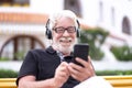 Portrait of happy senior white-haired man with beard sitting in a bench and listening to music smiling. Handsome people using Royalty Free Stock Photo