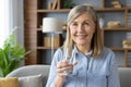 Portrait of happy senior woman in blue shirt looking at camera while raising hand with glass of still water. Positive Royalty Free Stock Photo