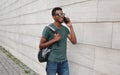 Portrait happy smiling african man calling on smartphone walking with backpack on city street over gray brick wall Royalty Free Stock Photo