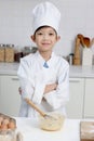 Portrait of happy smiling Asian boy in white chef uniform with hat standing with arms crossed and mixing eggs bowl on table at Royalty Free Stock Photo