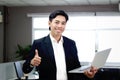 Portrait of happy smiling Asian businessman in black suit giving thumb up while holding laptop computer, standing inside office Royalty Free Stock Photo