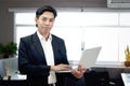 Portrait of happy smiling Asian businessman in black suit looking at the camera while holding laptop computer, standing inside Royalty Free Stock Photo