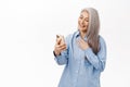 Portrait of happy smiling asian woman, old lady using smartphone app, holding mobile phone in hands, standing over white Royalty Free Stock Photo