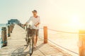 Portrait of a happy smiling barefoot man dressed in light summer clothes and sunglasses riding a bicycle on the wooden sea pier.