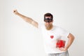 Portrait of happy smiling bearded male wears batman mask and casual white t shirt, holds wrapped gift box, raises hand