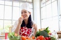 Portrait of happy smiling beautiful young Asian woman in red heart apron and chef hat holds and eats sliced carrot while sits Royalty Free Stock Photo