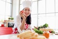 Portrait of happy smiling beautiful young Asian woman in red heart apron and chef hat holds red tomato while sits behind kitchen Royalty Free Stock Photo