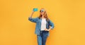 Portrait of happy smiling blonde young woman taking selfie with smartphone wearing denim jacket on yellow background Royalty Free Stock Photo