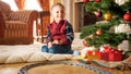 Portrait of happy smiling little boy sitting next to Christmas tree and presents from SAnta Claus at living room Royalty Free Stock Photo