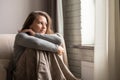 Portrait of happy smiling dreaming ÃÂaucasian young woman wrapped in a plaid sitting near window of apartments hugging knees. Royalty Free Stock Photo