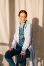 Portrait of happy smiling female doctor in white coat sitting on chair near window in sunny day in medical clinic office Royalty Free Stock Photo