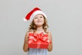 Portrait of a happy smiling little blonde girl on a gray background. A child in a Santa hat is holding a red crust with a gift. Royalty Free Stock Photo
