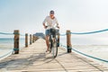 Portrait of a happy smiling man dressed in light summer clothes and sunglasses riding a bicycle on the wooden sea pier. Careless Royalty Free Stock Photo