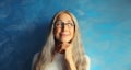 Portrait of happy smiling pensive middle-aged woman in eyeglasses looking away. Gray-haired female thinking on blue background Royalty Free Stock Photo