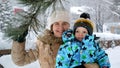Portrait of happy smiling toddler boy with mother enjoying falling snow from tree branch at park. Fun and joy on winter holidays, Royalty Free Stock Photo