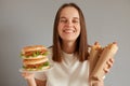 Portrait of happy smiling woman holding hot dog and burger wearing white T-shirt posing isolated over gray background, looking at Royalty Free Stock Photo