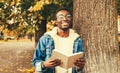 Portrait of happy smiling young african man student reading a book wearing eyeglasses in autumn city park Royalty Free Stock Photo