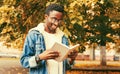 Portrait of happy smiling young african man student reading a book wearing eyeglasses in autumn city park Royalty Free Stock Photo