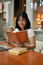 A portrait of a happy Asian woman enjoying coffee and reading a book in a beautiful cafe Royalty Free Stock Photo