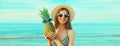 Portrait of happy smiling young woman with pineapple wearing straw hat on the beach on sea background at summer day Royalty Free Stock Photo