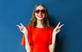 Portrait happy smiling young woman showing peace sign gesture wearing heart shaped sunglasses on blue wall Royalty Free Stock Photo