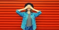 Portrait of happy surprised smiling young woman covering her eyes wearing black round hat, denim jacket on red background Royalty Free Stock Photo
