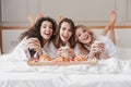 Portrait of happy three women 20s wearing bathrobe drinking latte and eating croissants in bed, during hen party in posh