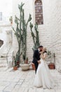 Happy wedding couple standing near ancient building, huge cactuses. Groom putting arm around brides neck, kissing.