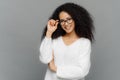 Portrait of happy woman with curly hair, kepes hands on frame of glasses, has gentle smile, wears white jumper, over grey